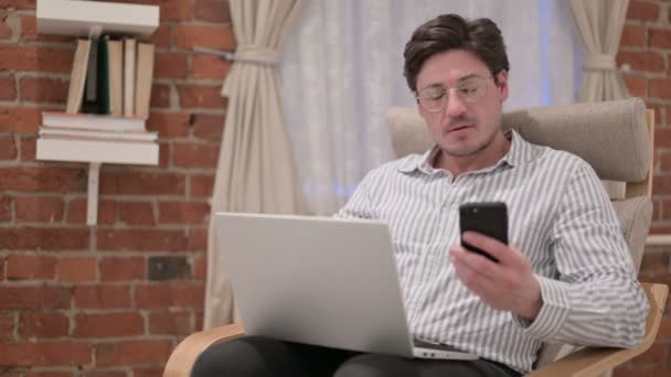 Hombre de mediana edad con ordenador portátil usando Smartphone en el sofá — Vídeos de Stock