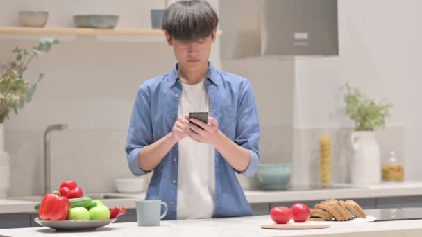 Asian Man using Smartphone while Standing in Kitchen — Stock Video