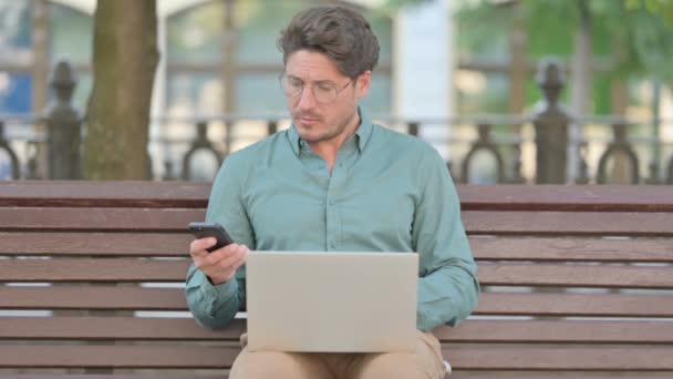 Чоловік середнього віку Перегляд на смартфоні під час роботи на ноутбуці — стокове відео