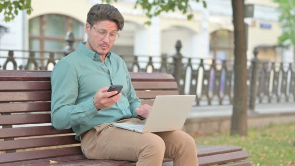 Ο άνθρωπος που περιηγείται στο Smartphone ενώ εργάζεται στο Laptop — Αρχείο Βίντεο