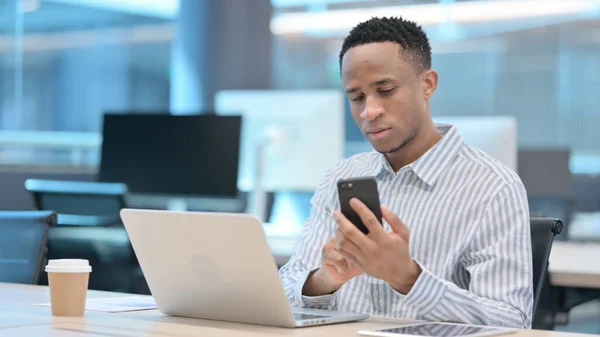 Afrikansk affärsman med bärbar dator med hjälp av smartphone på jobbet — Stockfoto