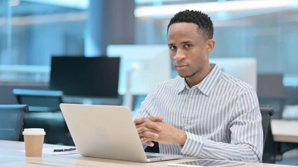 Empresário Africano Atraente Olhando para a câmera ao usar o laptop no escritório — Fotografia de Stock