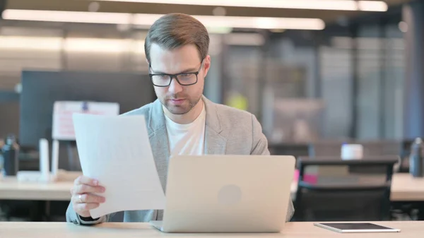 Jeune homme avec ordinateur portable lisant des documents au bureau — Photo