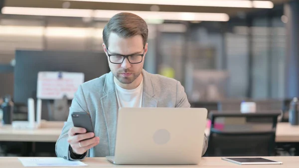 Νεαρός με Laptop Browsing Internet στο Smartphone στο Office — Φωτογραφία Αρχείου