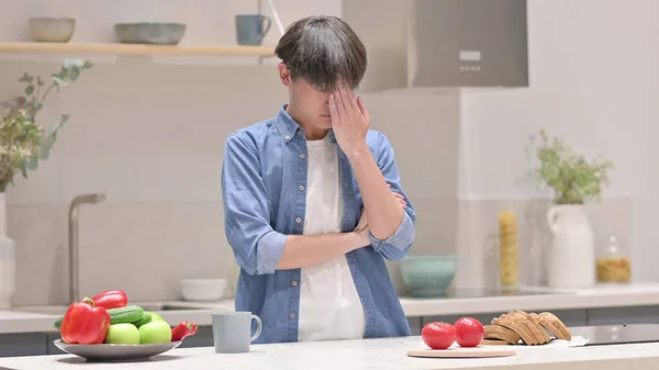 Serious Young Asian Man Thinking while Standing in Kitchen