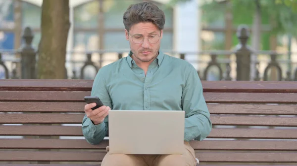 Ο άνθρωπος που περιηγείται στο Smartphone ενώ εργάζεται στο Laptop — Φωτογραφία Αρχείου