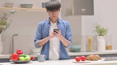 Genç Asyalı adam mutfakta dururken akıllı telefon kullanıyor.