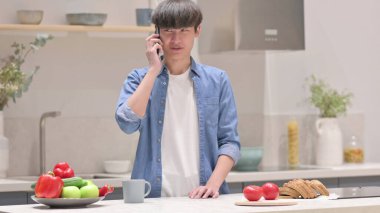 Genç Asyalı adam mutfakta dikilirken telefonda konuşuyor.