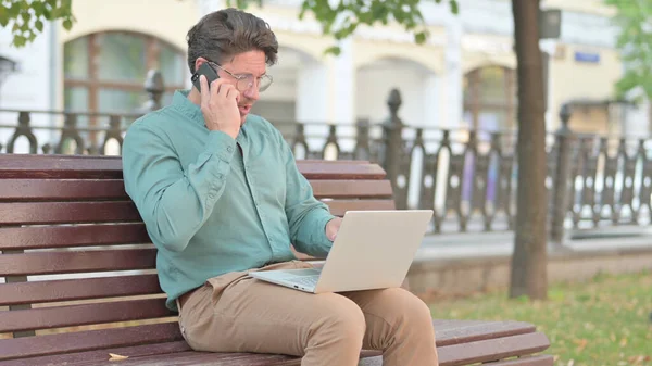 Starší dospělý muž mluví na telefonu při použití notebooku venkovní — Stock fotografie