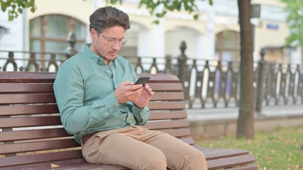Reifer erwachsener Mann surft auf Smartphone, während er auf Bank sitzt — Stockfoto