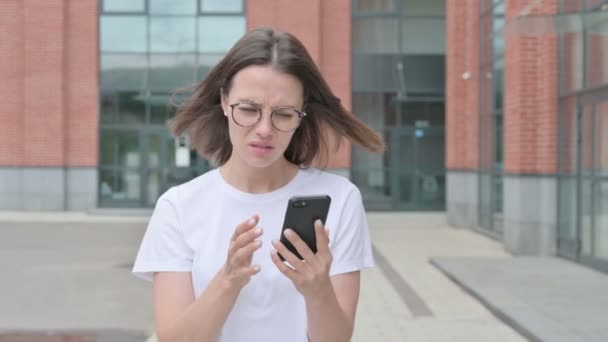 Молодая женщина реагирует на потерю на смартфоне во время прогулки по улице — стоковое видео