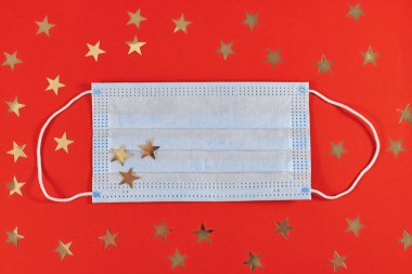 Corona Virüs Dönemi 'nde Noel, kırmızı arka planda altın yıldız süslemeleriyle çevrili tıbbi yüz maskesi ile kutlanır.