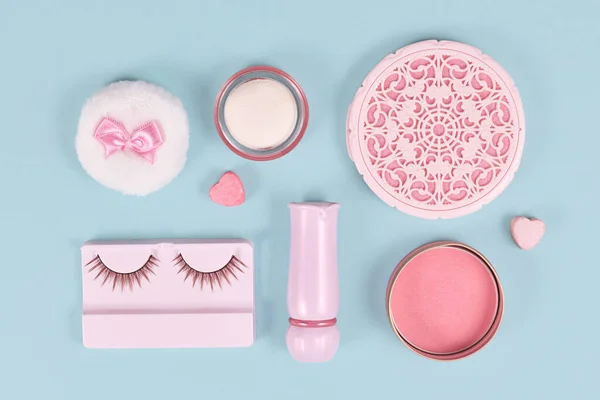 Pink makeup beauty products like fake lashes, blush, mirror or lipstick arranged on pastel blue background