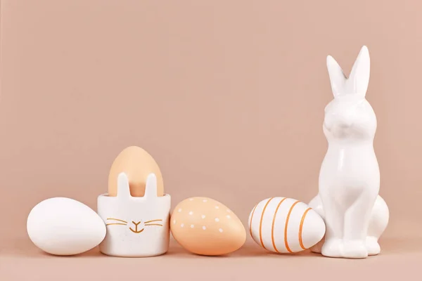 Natural colored Easter eggs with simple stripes and dots pattern with bunny shaped egg cup and sculpture beige background with empty copy space