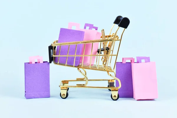 Golden shopping cart filled with pink and purple paper shopping bags on blue background