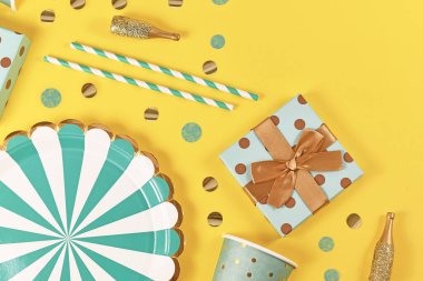 Teal mavisi kağıt tabaklar, hediye kutusu, küçük altın şampanya şişeleri ve sarı arka planda konfeti.