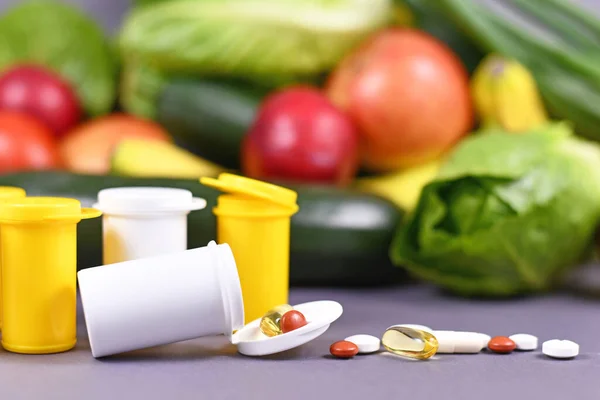Pills of food nutrition supplements spilling out of bottle in front of fruits and vegetables in background