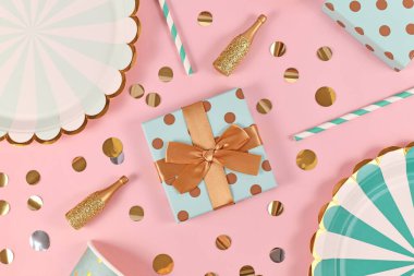 Üzerinde mavi kağıt tabaklar, küçük altın şampanya şişeleri ve pembe arka planda konfeti olan doğum günü hediyesi kutusu.