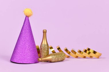 Parti şapkası, küçük altın şampanya şişeleri ve menekşe arkaplanda fotokopi alanı olan parti flamaları.