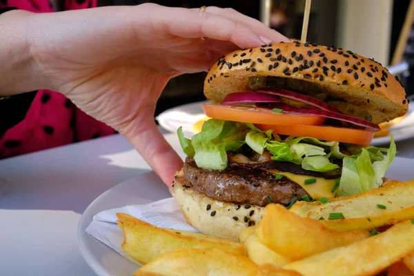 Mujer usando la mano para recoger una hamburguesa de ternera con papas fritas, tocino, lechuga, tomate, cebolla y encurtidos —  Fotos de Stock