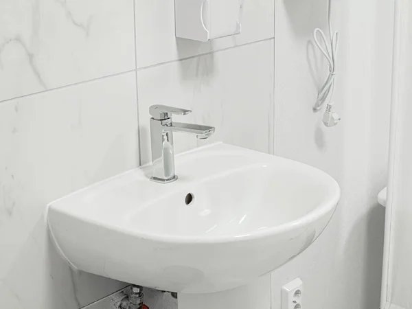 White new clean simple sink with metal shiny valve faucet in white interior of a medical clinic. Tiled wall in the background