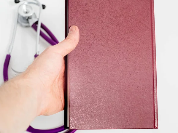 Doctor holding in hand red medical journal, work notebook, purple stethoscope on white background. Close-up.