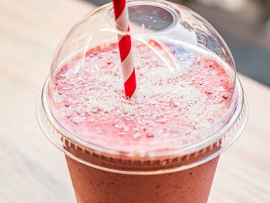 Plastik bardakta pembe meyve kokteyli ve beyaz-kırmızı spiral tüp ile milkshake sokak kafesindeki masada duruyor. Makro