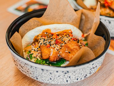 Beyaz tabakta tavuklu bao. Etli, salatalı, sebzeli, soslu, susam tohumlu pirinç çöreği. Bir kafede, restoranda fast food.