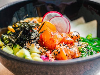 Somon, avokado, turp, mısır, yosun, bezelye, nori, daikon, susam tohumu. Kafede, restoranda, ahşap masada siyah tabakta Asya yemeği. Yakın plan.