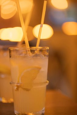 Limonata ve buzlu dilimlenmiş limon dilimleri. Kafedeki masada iki pipetle serinletici soğuk bir içecek. Bulanık sıcak arkaplan
