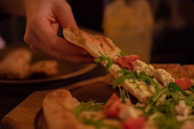 Sıcak erimiş peynir, bir İtalyan restoranında sıcak bir akşam ışığında yemek için bir kadın tarafından domates ve roka ile bir dilim pizzanın üzerine gerilir. Yakın plan.