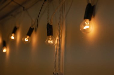 Yuvarlak küçük ampullerden oluşan bir çelenk. Siyah tabanlı. Ilık turuncu ışık, karanlık bir odada beyaz bir duvarda asılı. Çatı katı tarzı dekor. Yakın plan.