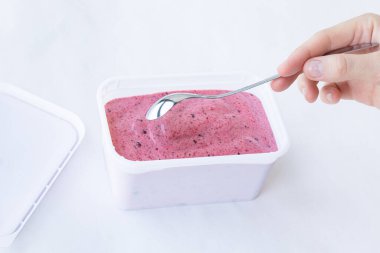 Beyaz arka planda elinde metal gümüş kaşıkla beyaz kapağı olan dikdörtgen beyaz bir kabın içinde siyah üzümlü dondurma pembe renginde krema dokusu. Üst Manzara. Yeme konsepti