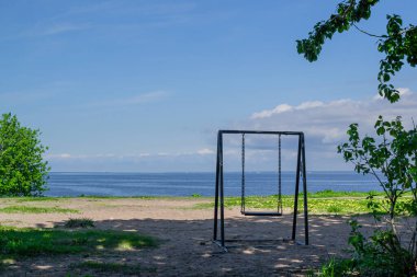 Boş bir salıncak, bir ağacın gölgesinde duruyor. Yeşil çimenlerin arasında ve güneşin ışığında. Mavi denizin ve gökyüzünün arka planında bulutlarla.