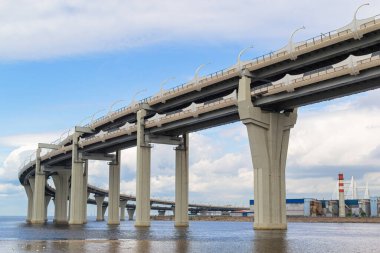 Büyük yuvarlak, kontrollü otoyol köprüsü bulutlu mavi gökyüzünün arka planına karşı şehrin kıyısı yakınlarındaki deniz koyunun üzerinden geçen yol, endüstri. Saint Petersburg, Avrupa