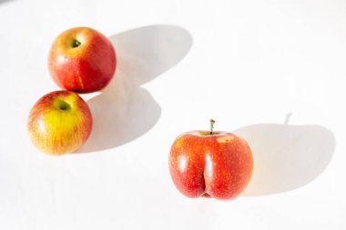 Bright ripe red apple of an unusual shape of an ass in the light of the sun on a white background with two usual apples with long shadows. Top view. Nonconformism, body positive, dissimilarity, uniqueness concepts