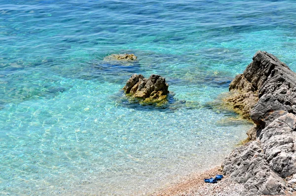 Plage rocheuse avec eau bleue claire et pantoufles bleues par une journée ensoleillée — Photo