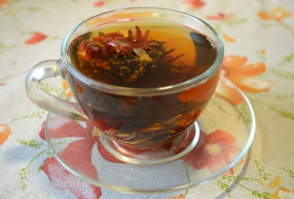 Blütentee in der durchsichtigen Tasse auf transparentem Untersetzer und auf Tischdecke mit Blumen — Stockfoto