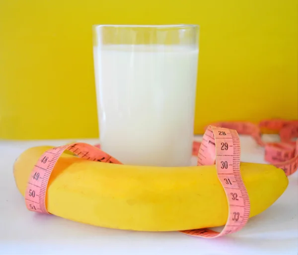 Un vaso de leche, un plátano y una cinta métrica sobre un fondo amarillo. Concepto de dieta — Foto de Stock