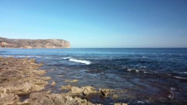 Akdeniz manzarası, kıyıya vuran küçük dalgalar ve arkasında deniz feneri olan bir pelerin.