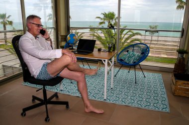 Adam ev ofisinden deniz manzaralı şortla ofis işi yapıyor.