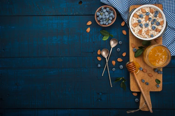 Um pequeno-almoço saudável em um fundo de madeira azul escuro: Aveia, leite, mirtilos, mel e amêndoas. Estilo rústico . — Fotografia de Stock
