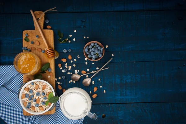 Un mic dejun sănătos pe un fundal de lemn albastru închis: fulgi de ovăz, lapte, afine, miere și migdale. Stilul rustic . — Fotografie, imagine de stoc