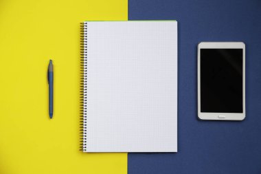 Açık bir defterin üst görünümü, bir tablet bilgisayar ve mavi ve sarı arka gruba karşı bir kalem