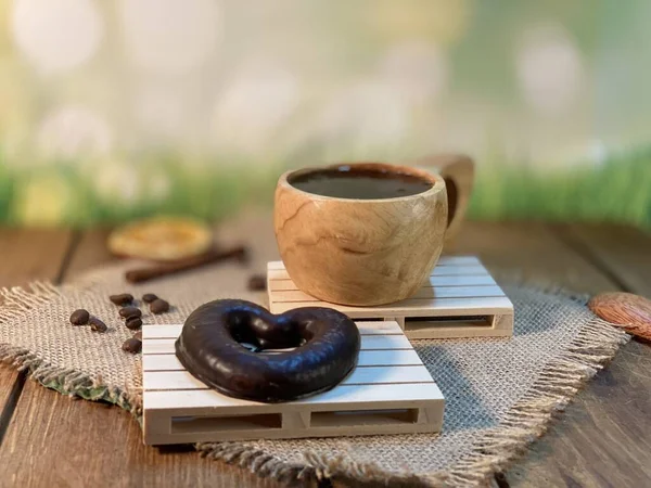 Oak table with visible grains, stand, pallet, on it there is a cup made of wood with black coffee. Coffee beans scattered on a jute napkin, a wooden spoon, background green grass and gingerbread heart is lying on a wooden pallet