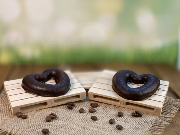 Oak table with visible rings on the stand. A palette on it stands a cup made of wood with black coffee. On a jute napkin, coffee beans are scattered, a wooden spoon, the background green grass and gingerbread hearts lie on a wooden pallet