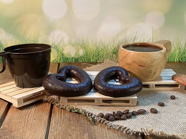 Oak table with visible grains, birch stand, on it a cup made of wood with black coffee. Coffee beans scattered on a jute napkin, a wooden spoon, background green grass and gingerbread heart is lying on the palette
