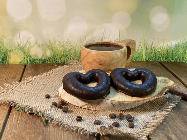 Oak table with visible grains, birch stand, on it a cup made of wood with black coffee. Coffee beans scattered on a jute napkin, a wooden spoon, background green grass and gingerbread heart is lying on the palette