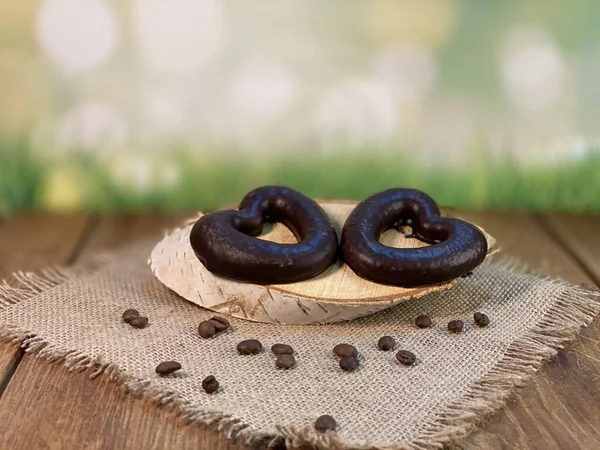 Oak table with visible grains, birch stand, on it a cup made of wood with black coffee. Coffee beans scattered on a jute napkin, a wooden spoon, background green grass and gingerbread heart is lying on the palette