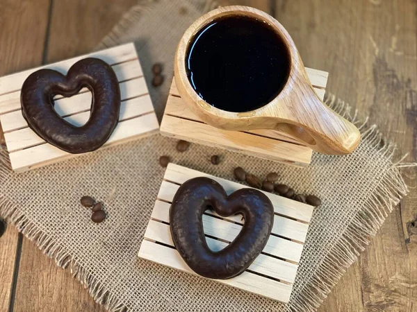 An oak table with visible grains stands a cup made of wood with black coffee. On a jute napkin, the coffee beans are scattered and the gingerbread hearts lie on the pallets